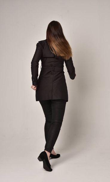 full length portrait of a brunette girl wearing long black coat, standing pose with back to the camera on grey studio background. - Фото, изображение