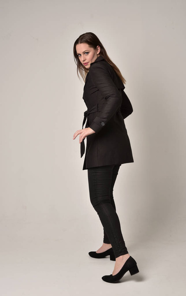 портрет брюнетки в длинном черном пальто, стоящей в позе спиной к камере на сером фоне студии
. - Фото, изображение