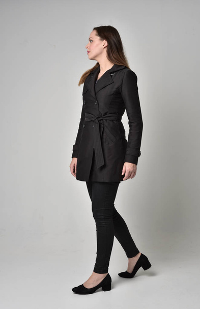 full length portrait of a brunette girl wearing long black coat, standing pose in side profile on grey studio background. - Foto, Bild