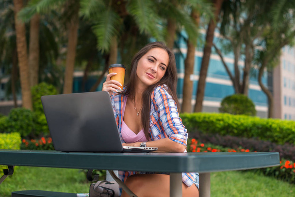 Mujer joven tomando un descanso para relajarse del trabajo en el portátil en el parque mientras sostiene la taza de café en una mano con los ojos cerrados y sonreír placer
 - Foto, imagen