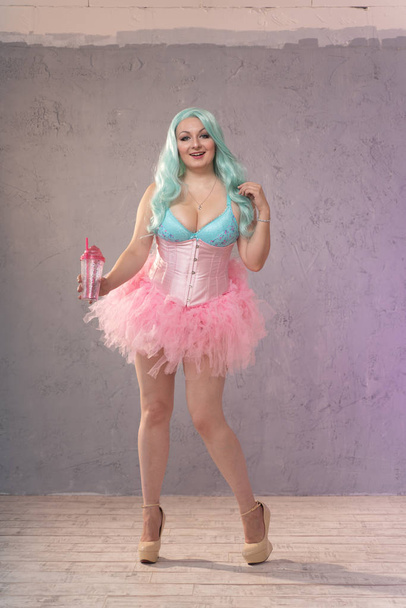 chubby blonde girl in pink lingerie, corset and tutu skirt posing with a glass of water on grey background - Valokuva, kuva