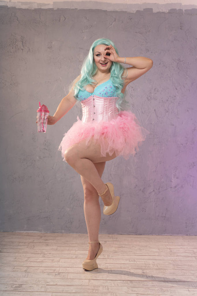пухленькая блондинка в розовом нижнем белье, корсет и юбка в пачке позирует со стаканом воды на сером фоне
 - Фото, изображение