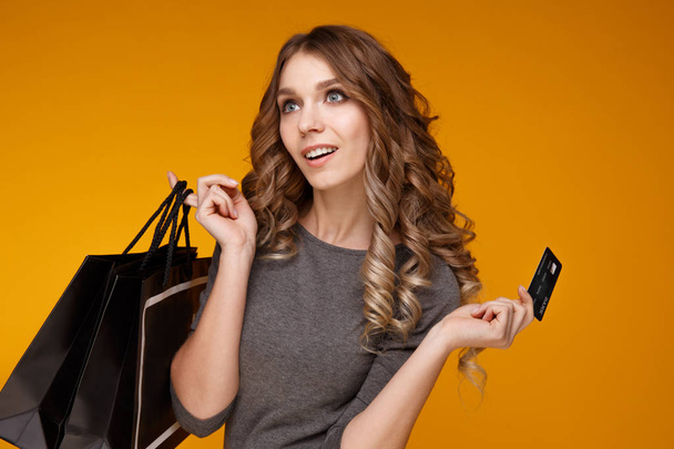 ztratit up portrét šťastná mladá bruneta žena držící kreditku a barevné nákupní tašky, při pohledu na fotoaparát, izolované na žlutém podkladu - Fotografie, Obrázek
