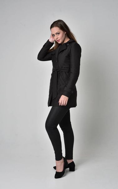 full length portrait of a brunette girl wearing long black coat, standing pose in side profile on grey studio background. - Photo, Image