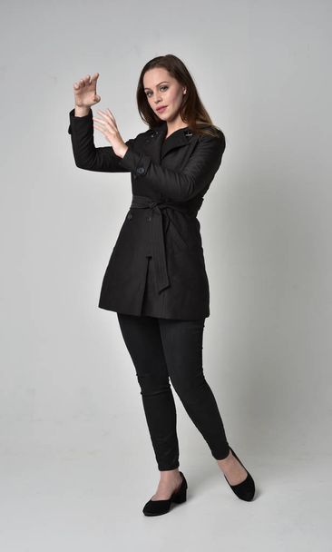 Pełna długość Portret brunetka Girl ubrany długi czarny płaszcz, stojący poza w Profil boczny na studio szary tło. - Zdjęcie, obraz