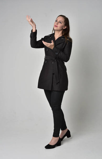 full length portrait of a brunette girl wearing long black coat, standing pose in side profile on grey studio background. - Фото, изображение