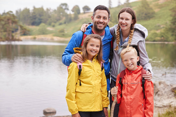 Young family standing on the shore of a lake in the countryside looking to camera smiling, Lake District, UK - 写真・画像