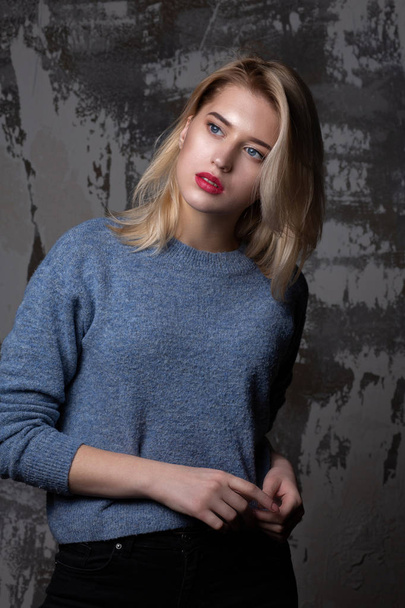 Fashionable blonde woman with lush hair wearing blue sweater, posing in studio with shadows - Фото, зображення