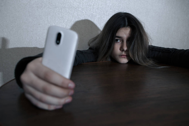 Soltera adolescente triste sosteniendo un teléfono móvil lamentándose sentada en la cama en su dormitorio con una luz oscura
 - Foto, Imagen