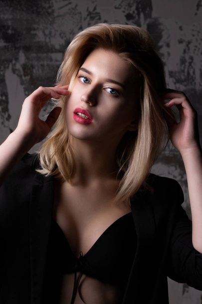 attraktive blonde Frau mit perfekter Haut trägt schwarze Jacke und BH posiert im Studio mit Schatten - Foto, Bild