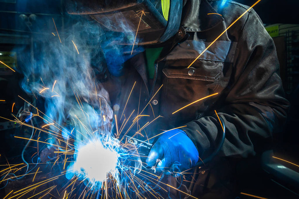 Man welder in welding mask, building uniform and blue protective gloves welds metal car muffler with welding machine in auto repair shop, in the background construction site - Photo, Image