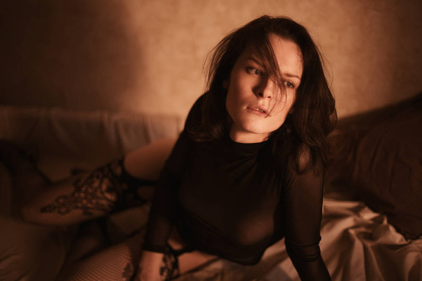attractive young woman with a big beautiful breasts half lying in a bed naked or nude in a seductive fishnet stockings and transparent lace body under worm sunset light. - Fotografie, Obrázek