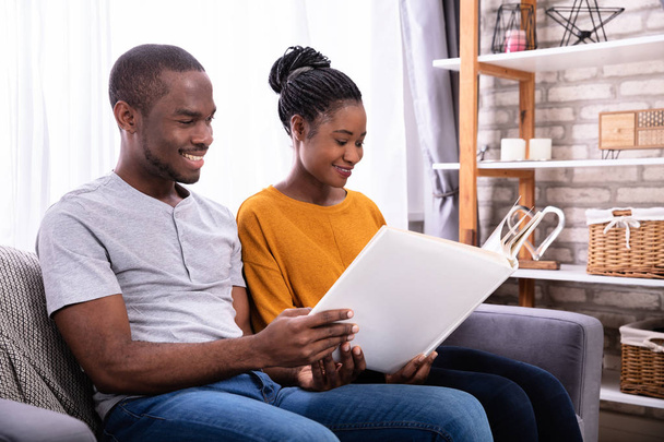 Smiling Young African Couple Sitting On Sofa Looking At Photo Album At Home - Φωτογραφία, εικόνα