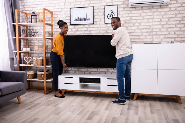 Vista lateral de la joven pareja africana que levanta la televisión en casa
 - Foto, Imagen