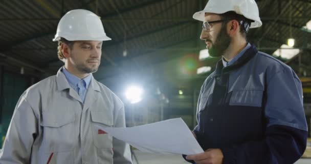 Factory worker and engineer in helmets with big paper drawings shaking hands and agreed on the project. Inside a big manufactory. Portrait shot. Close up - Footage, Video