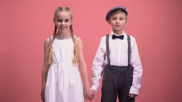 Smiling little boy and girl looking into camera holding hands on pink background - Filmagem, Vídeo