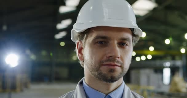 Lähikuva houkutteleva valkoihoinen insinööri kypärä kääntää päänsä kameraan ja hymyilee. Iso tehdastalo hämärtyneellä taustalla. Muotokuva - Materiaali, video