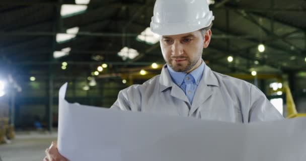 Portrait de l'homme caucasien attrayant, ingénieur dans un casque tenant un grand papier avec des dessins dans ses mains et en regardant attentivement. La grande usine sur le fond. Gros plan
 - Séquence, vidéo