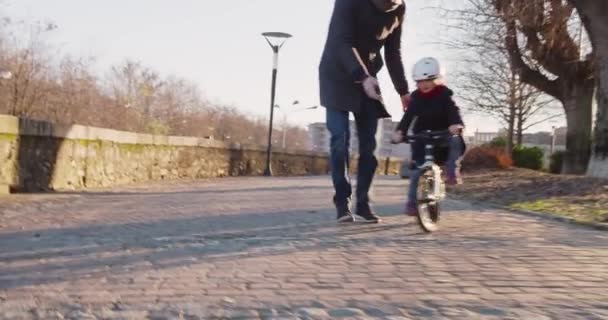 Daughter child girl learning riding bycicle with dad teaching in city.Growing,childhood,active safety family.Sidewalk urban outdoor.Warm sunset cold weather backlight.4k slow motion 60p front video - Filmagem, Vídeo