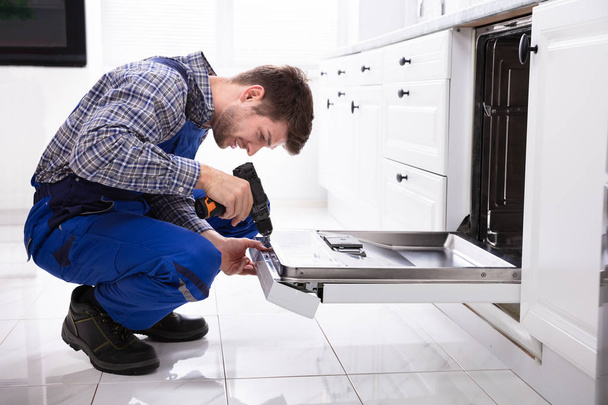 Young Repairman Fixing Dishwasher With Electric Drill In Kitchen - Photo, image