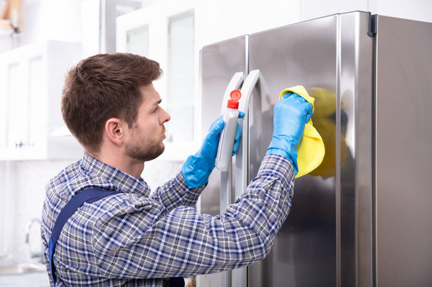 Side View Of Young Man Cleaning Refrigerator With Napkin And Bottle - Foto, imagen