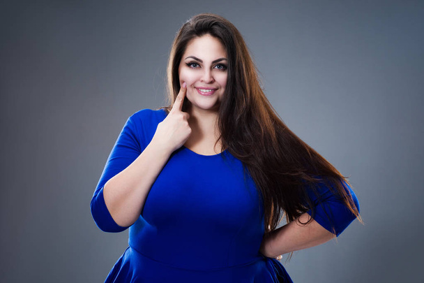 Zahngesundheitskonzept, dicke Frau zeigt auf ihre Zähne, Plus-Size-Model im blauen Kleid auf grauem Studiohintergrund - Foto, Bild