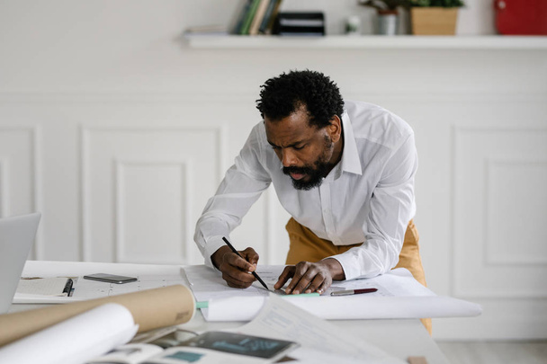 Serious African man architect drawing on blueprints at his office. - Photo, image
