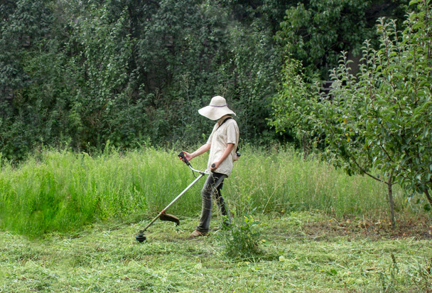 Střih trávníku. Dívka v pracovní oděvy seká trávu na zahradě. Velký klobouk a brýle chránit dívku. Holky mění stereotypy. Koncept silných žen - Fotografie, Obrázek