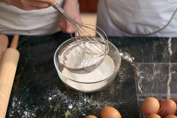 Крупный план неузнаваемой женщины, держащей решето с мукой и готовящейся взбивать его яйцами во время приготовления теста для печенья
 - Фото, изображение