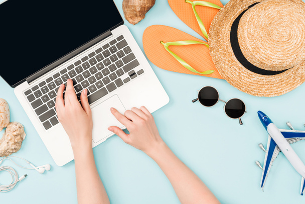cropped view of woman using laptop near earphones, sunglasses, seashells, flip flops, toy plane and straw hat on blue background - Photo, Image