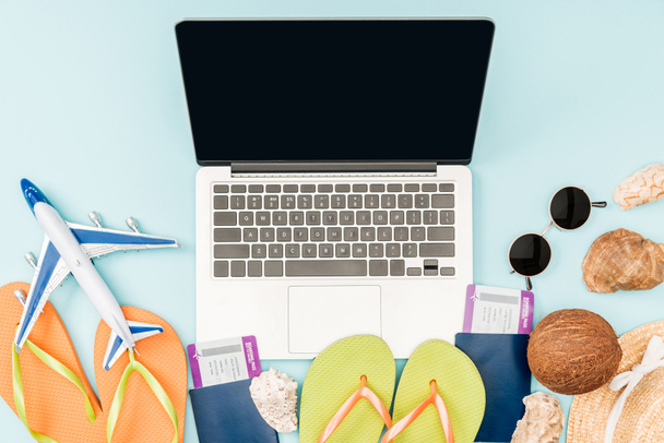 top view of laptop with blank screen, flip flops, passports with air tickets, sunglasses, seashells, straw hat and plane toy on blue background - Foto, Imagen