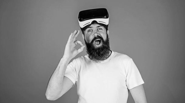 Man with long beard and funny face showing OK gesture. Hipster with trendy beard wearing VR goggles, digital technologies concept. Bearded man testing 3D video plugin isolated on blue background - Фото, изображение