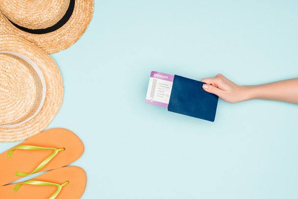 cropped view of woman holding air ticket and passport near flip flops and straw hats on blue background - Photo, Image