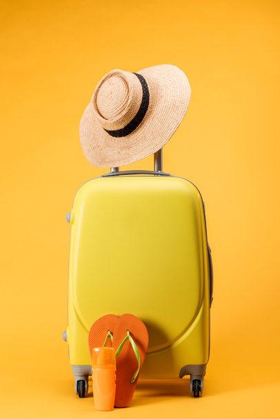 bolsa de viaje con ruedas, sombrero de paja y chanclas sobre fondo amarillo
 - Foto, Imagen