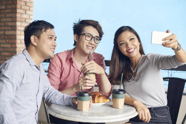 Szczęśliwy nowoczesne Azjatyckie kolegów z kawę i bułeczki w kawiarni biorąc selfie z telefonu - Zdjęcie, obraz