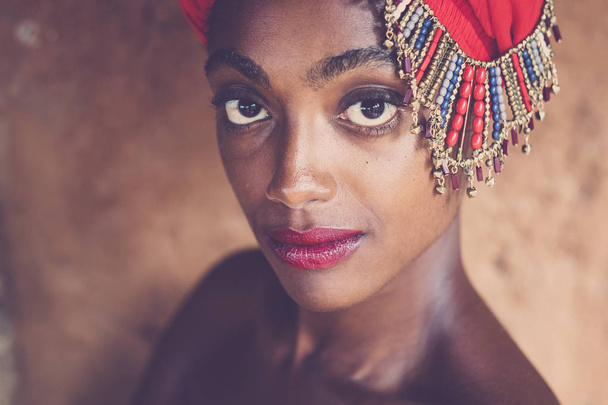 Ομορφιά και όμορφο μαύρο αφρικανικό κορίτσι με παραδοσιακό καπέλο μόδας με κοσμήματα που θέτουν και κοιτάζοντας την κάμερα με μεγάλα υπέροχα μάτια  - Φωτογραφία, εικόνα