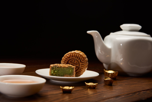 tradicionales deliciosos pasteles de luna chinos, tetera y lingotes de oro en mesa de madera aislada en negro
 - Foto, Imagen