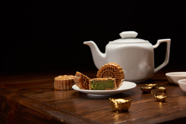 deliciosos bolos chineses tradicionais, panela de chá e lingotes de ouro na mesa de madeira isolada em preto
 - Foto, Imagem