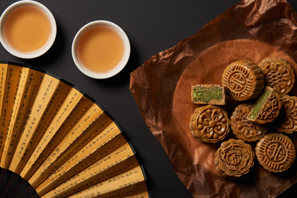 vista superior de mooncakes chineses tradicionais com chá e ventilador de mão isolado em preto
 - Foto, Imagem