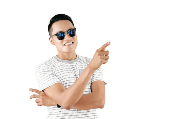 Taille-up portret van jonge Aziatisch vent in stijlvolle zonnebril tonen wijsvinger gebaar en lacht vrolijk op witte achtergrond - Foto, afbeelding