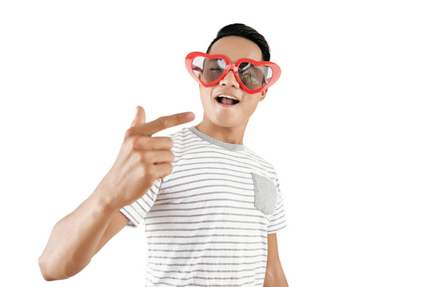 Portrait of young handsome Asian man wearing trendy heart-shaped sunglasses posing on camera against white background - Photo, Image
