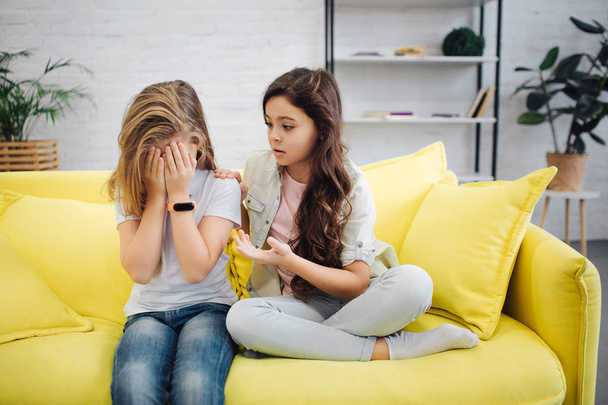Upset girl on left hide face with hands. She cry. Teenager sit with her friend on yellow sofa in room. Brunette try to comfort her. - Фото, зображення