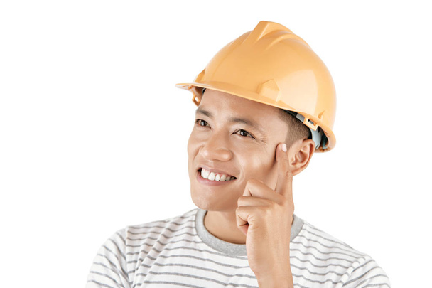 Portrait of young Asian man wearing hard hat holding finger on temple while generating ideas and smiling happily against white background - Foto, afbeelding