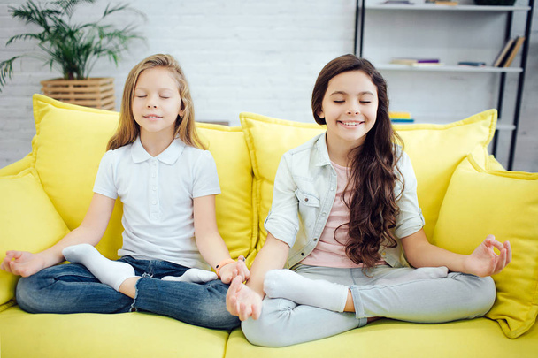 Two young teenagers sit in lotus pose on yellow sofa in room. They meditate. Girls express positive emotions. They keep eyes closed. - Zdjęcie, obraz
