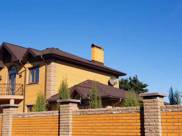 Casa de tijolo grande com chaminé, sarjeta de chuva e cerca de tijolo sob céu azul claro, vista lateral
 - Foto, Imagem