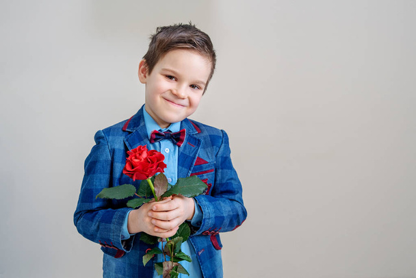 Μικρό αγόρι στο κοστούμι στέκεται με κόκκινο τριαντάφυλλο, σε ανοιχτόχρωμο φόντο, st. ημέρα του Αγίου Βαλεντίνου ημέρα έννοια - Φωτογραφία, εικόνα