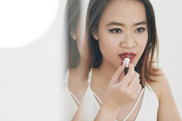 hübsche junge Vietnamesin trägt roten Lippenstift vor dem Spiegel auf - Foto, Bild