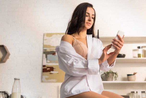 hermosa mujer en camisa blanca usando teléfono inteligente en la mañana en casa
 - Foto, Imagen