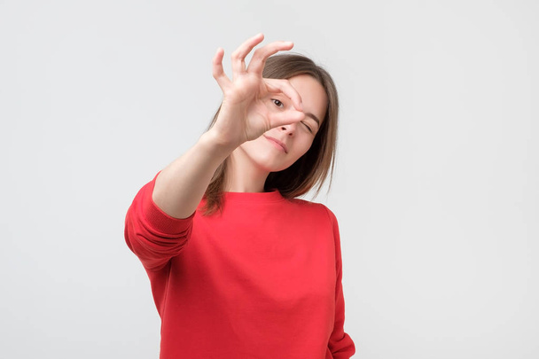 Young adult woman wearing red sweater doing ok gesture smiling, eye looking through fingers. Unbelieving expression. - Fotografie, Obrázek