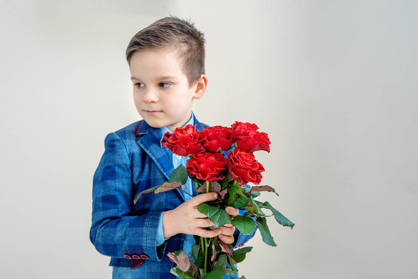 Adorable little boy in suit with a bouquet of red roses in hands on a light background, st. valentine's day concept - 写真・画像